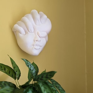 May include: A white plaster wall sculpture of a face with closed eyes and a hand covering the forehead. The sculpture is mounted on a yellow wall, with green leafy plants in the foreground.