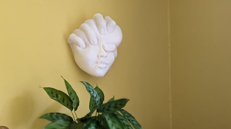 May include: A white plaster wall sculpture of a face with closed eyes and a hand covering the forehead. The sculpture is mounted on a yellow wall, with green leafy plants in the foreground.