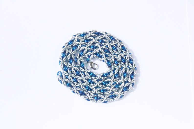 Puede incluir: Un collar enrollado con un dise&ntilde;o de cota de malla. El collar presenta eslabones plateados y azules alternados, creando un contraste llamativo. Un cierre plateado asegura los extremos. La joya se muestra sobre un fondo blanco.