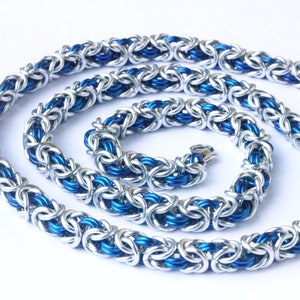 Puede incluir: Un collar de cota de malla plateado y azul. El collar est&aacute; hecho de anillos de metal entrelazados, con un exterior plateado y un interior azul vibrante. La cadena est&aacute; dispuesta en espiral sobre un fondo blanco. El collar tiene un cierre.