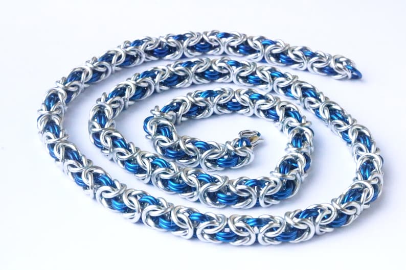 Puede incluir: Un collar de cota de malla plateado y azul. El collar est&aacute; hecho de anillos de metal entrelazados, con un exterior plateado y un interior azul vibrante. La cadena est&aacute; dispuesta en espiral sobre un fondo blanco. El collar tiene un cierre.