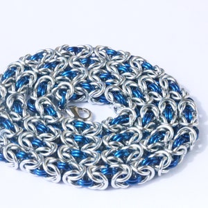 Puede incluir: Un collar de cota de malla plateado y azul con cierre de mosquet&oacute;n. La cota de malla est&aacute; hecha de anillos de metal entrelazados, con anillos plateados y azules alternados que crean un patr&oacute;n texturizado. El collar se muestra sobre un fondo blanco.