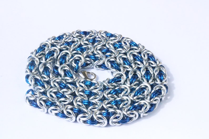 Puede incluir: Un collar de cota de malla plateado y azul con cierre de mosquet&oacute;n. La cota de malla est&aacute; hecha de anillos de metal entrelazados, con anillos plateados y azules alternados que crean un patr&oacute;n texturizado. El collar se muestra sobre un fondo blanco.