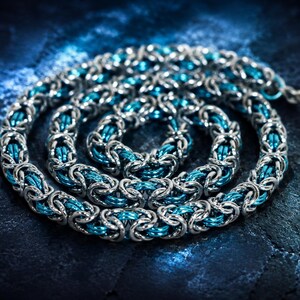 Puede incluir: Un collar de cota de malla plateado con detalles en azul turquesa, enrollado sobre una superficie texturizada azul oscuro. El intrincado dise&ntilde;o presenta anillos de metal entrelazados, creando una textura visual llamativa. Esta joya artesanal es adecuada para diversas ocasiones.
