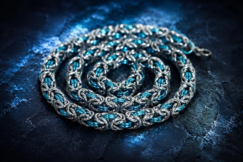 Puede incluir: Un collar de cota de malla plateado con detalles en azul turquesa, enrollado sobre una superficie texturizada azul oscuro. El intrincado dise&ntilde;o presenta anillos de metal entrelazados, creando una textura visual llamativa. Esta joya artesanal es adecuada para diversas ocasiones.