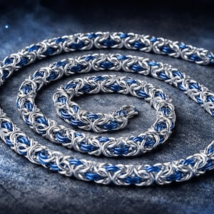Puede incluir: Un collar de cadena plateada con detalles azules, enrollado en forma de espiral. La cadena est&aacute; hecha de eslabones met&aacute;licos entrelazados, creando una superficie texturizada. El collar se muestra sobre un fondo oscuro y texturizado.