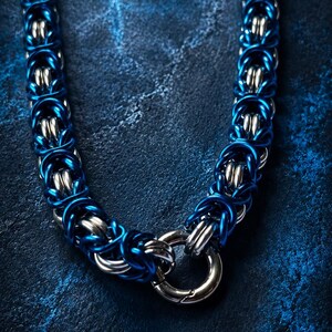 Puede incluir: Primer plano de un collar de cota de malla azul y plateado. El collar est&aacute; hecho de anillos de metal entrelazados, con un cierre en la parte inferior. El fondo es una superficie texturizada azul oscuro. Aproximadamente 40 cm de largo.