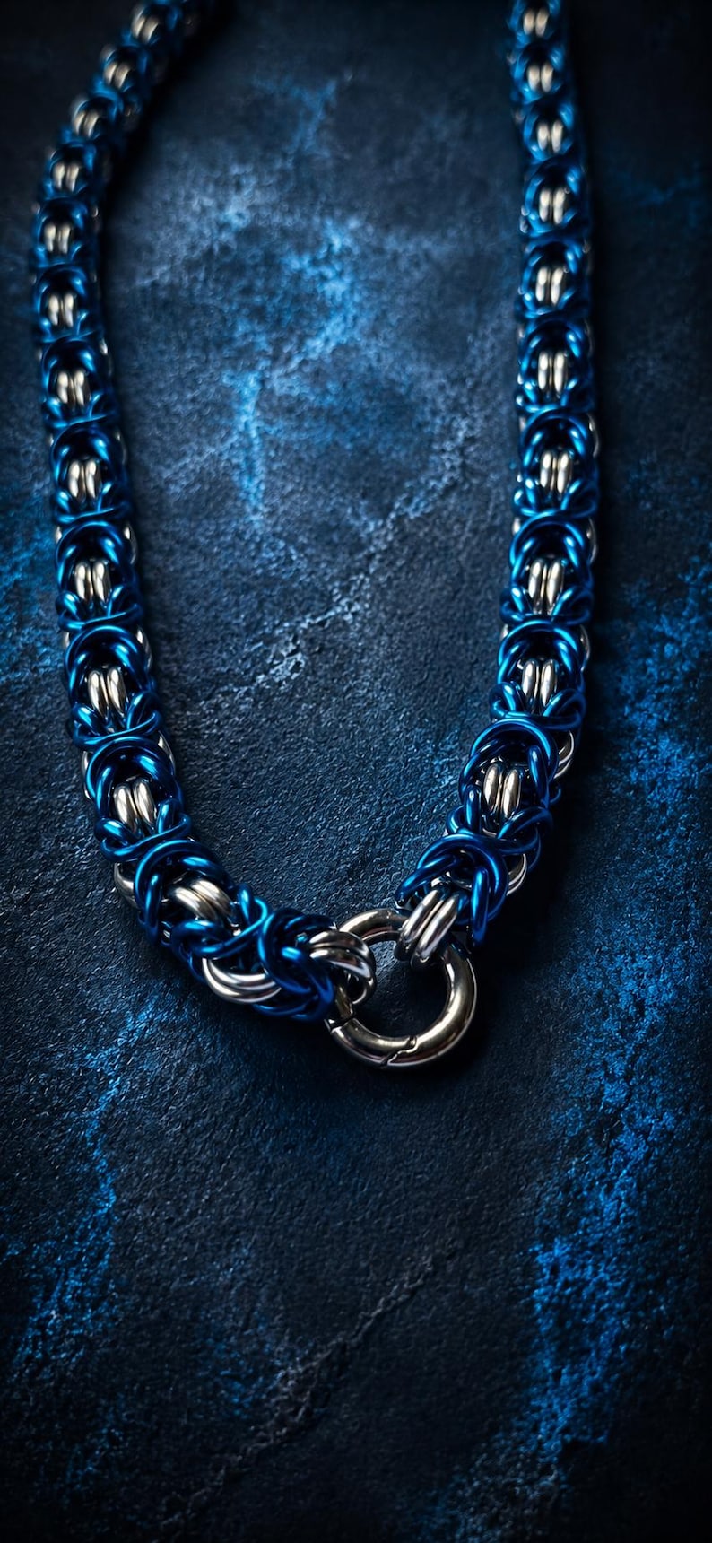 Puede incluir: Primer plano de un collar de cota de malla azul y plateado. El collar est&aacute; hecho de anillos de metal entrelazados, con un cierre en la parte inferior. El fondo es una superficie texturizada azul oscuro. Aproximadamente 40 cm de largo.