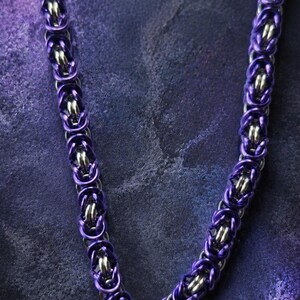 Puede incluir: Un collar de cota de malla morado y plateado. El collar est&aacute; hecho de anillos de metal entrelazados, con un vibrante tono morado en los anillos exteriores y anillos interiores plateados. El collar tiene un cierre circular.