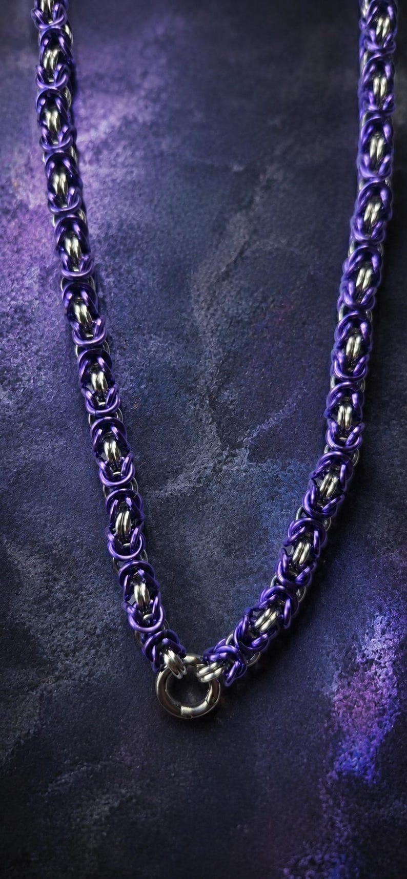 Puede incluir: Un collar de cota de malla morado y plateado. El collar est&aacute; hecho de anillos de metal entrelazados, con un vibrante tono morado en los anillos exteriores y anillos interiores plateados. El collar tiene un cierre circular.