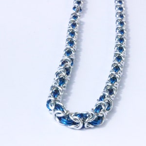 Puede incluir: Un collar de cota de malla plateado con detalles azules. El collar est&aacute; hecho de anillos de metal entrelazados, creando una apariencia texturizada y detallada. El color azul es visible dentro de los eslabones, agregando un toque de color a la joya.