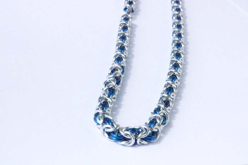 Puede incluir: Un collar de cota de malla plateado con detalles azules. El collar est&aacute; hecho de anillos de metal entrelazados, creando una apariencia texturizada y detallada. El color azul es visible dentro de los eslabones, agregando un toque de color a la joya.