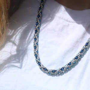 Puede incluir: Un collar de cadena plateada con detalles azules. El collar presenta un dise&ntilde;o tejido hecho de anillos de metal entrelazados. Los elementos azules est&aacute;n espaciados a lo largo de la cadena, proporcionando un color contrastante.
