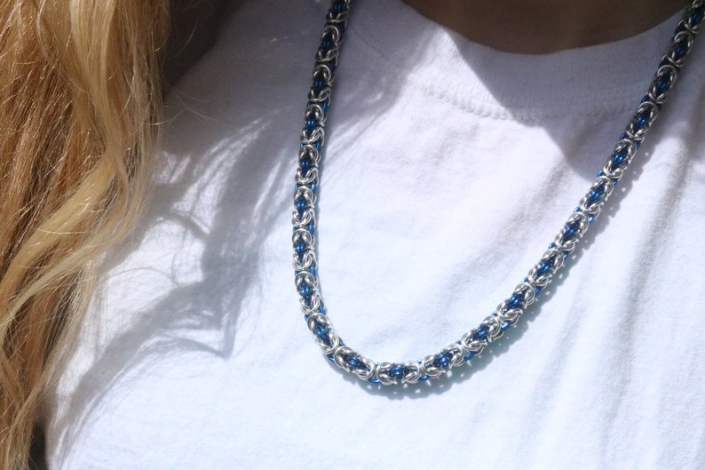 Puede incluir: Un collar de cadena plateada con detalles azules. El collar presenta un dise&ntilde;o tejido hecho de anillos de metal entrelazados. Los elementos azules est&aacute;n espaciados a lo largo de la cadena, proporcionando un color contrastante.