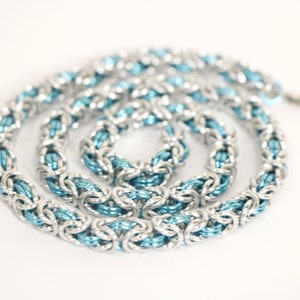 Puede incluir: Un collar de cota de malla plateado con un n&uacute;cleo interno azul claro. El collar est&aacute; hecho de anillos de metal entrelazados, creando una apariencia texturizada y tejida. La cadena est&aacute; dispuesta en espiral sobre un fondo blanco.