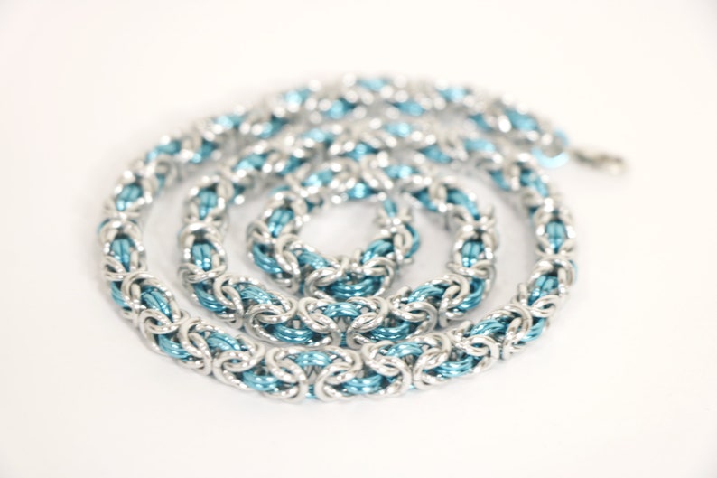 Puede incluir: Un collar de cota de malla plateado con un n&uacute;cleo interno azul claro. El collar est&aacute; hecho de anillos de metal entrelazados, creando una apariencia texturizada y tejida. La cadena est&aacute; dispuesta en espiral sobre un fondo blanco.
