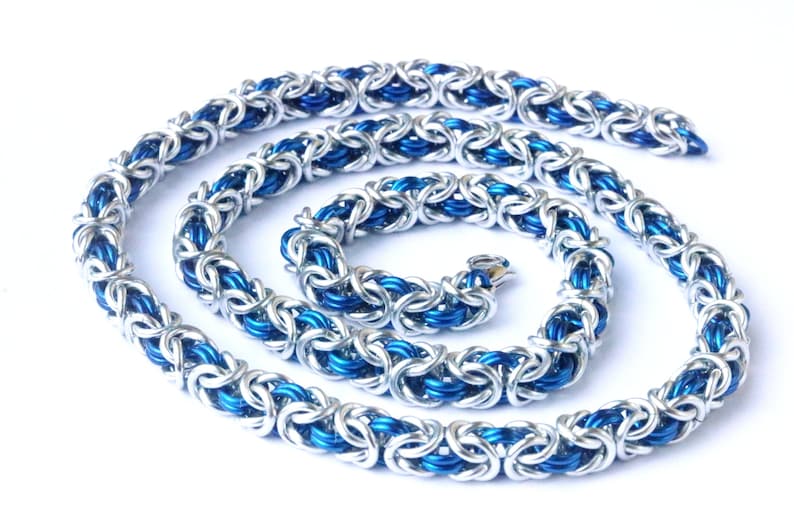 Puede incluir: Un collar de cota de malla plateado y azul, enrollado sobre una superficie blanca. El collar est&aacute; hecho de anillos de metal entrelazados, con un azul brillante en el centro y plata en el exterior.