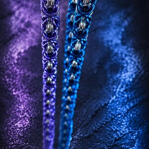 Puede incluir: Primer plano de dos collares de cota de malla, uno en morado oscuro y otro en azul. Cada collar presenta una serie de anillos met&aacute;licos entrelazados, con un anillo m&aacute;s grande en la parte superior. El fondo es una superficie oscura y texturizada con iluminaci&oacute;n p&uacute;rpura y azul.