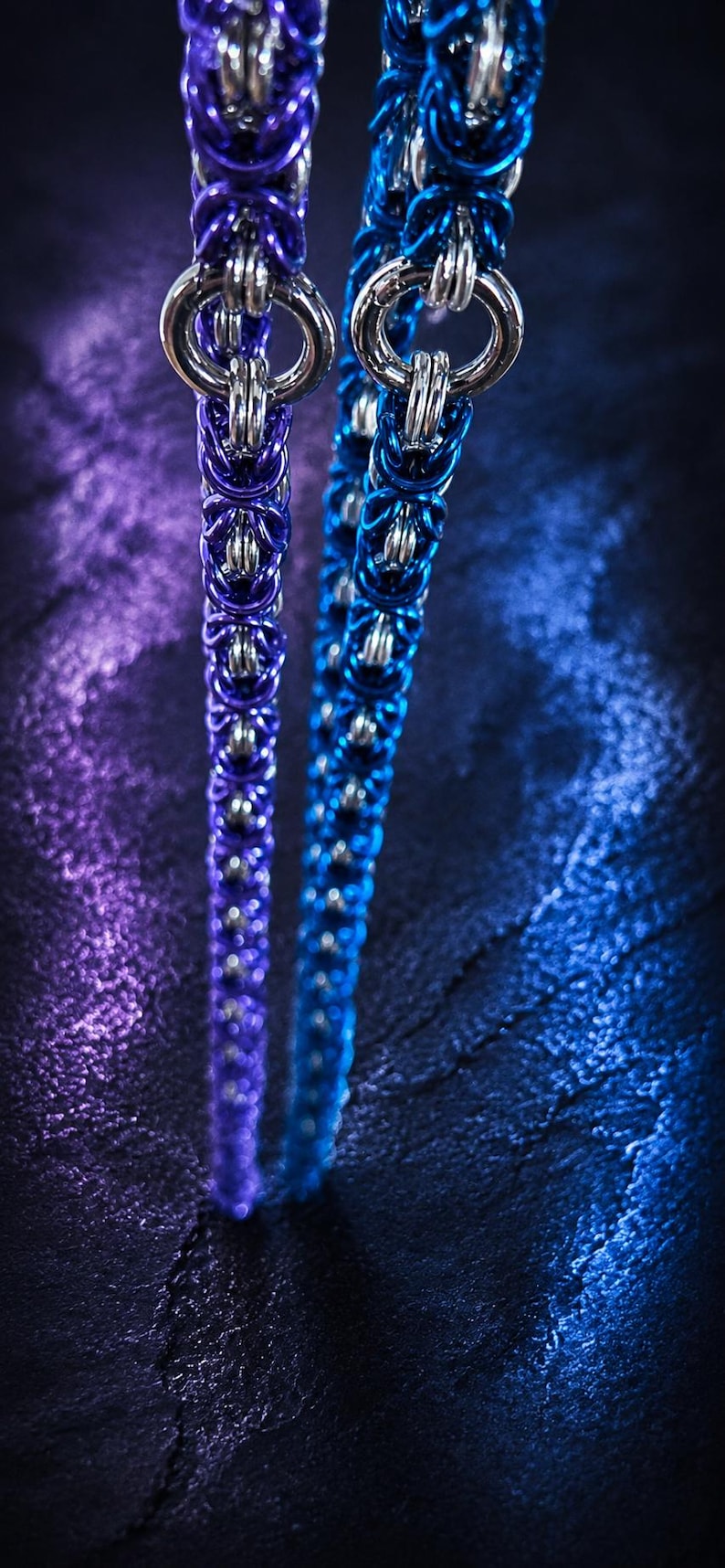 Puede incluir: Primer plano de dos collares de cota de malla, uno en morado oscuro y otro en azul. Cada collar presenta una serie de anillos met&aacute;licos entrelazados, con un anillo m&aacute;s grande en la parte superior. El fondo es una superficie oscura y texturizada con iluminaci&oacute;n p&uacute;rpura y azul.