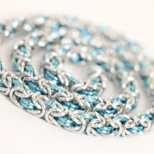 Puede incluir: Un collar de cota de malla plateado con un n&uacute;cleo interno azul claro. La cadena est&aacute; hecha de anillos de metal entrelazados, creando una apariencia texturizada y tejida. El collar se muestra sobre un fondo blanco.
