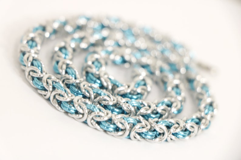 Puede incluir: Un collar de cota de malla plateado con un n&uacute;cleo interno azul claro. La cadena est&aacute; hecha de anillos de metal entrelazados, creando una apariencia texturizada y tejida. El collar se muestra sobre un fondo blanco.