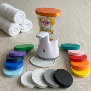 Könnte beinhalten: Ein weißer automatischer Seifenspender mit orangefarbenem Deckel und Sagrotan-Etikett, umgeben von einer Sammlung kreisförmiger, mehrfarbiger Untersetzer in den Farben Grün, Lila, Blau, Weiß, Grau, Schwarz, Rosa, Rot, Orange und Gelb.