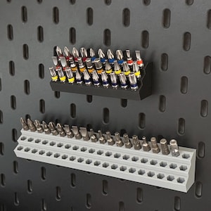 Könnte beinhalten: Zwei gestufte Aufbewahrungsregale für Schraubendreher-Bits, montiert an einer dunkelgrauen Lochwand. Das obere schwarze Regal enthält Metall-Bits mit farbigen Ringen. Das untere hellgraue Regal verfügt über leere, sechseckige Steckplätze.
