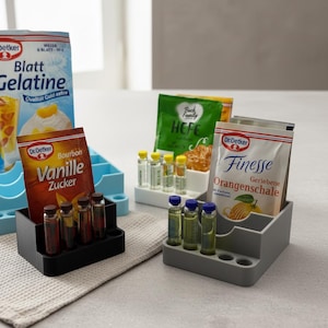 Könnte beinhalten: Drei gestufte Kunststoff-Organizer in Schwarz, Weiß und Hellblau, die jeweils kleine Glasfläschchen und verschiedene Backzutaten-Tüten enthalten, darunter Dr. Oetker Blatt Gelatine, Vanille Zucker, Hefe und Finesse Geriebene Orangenschale.