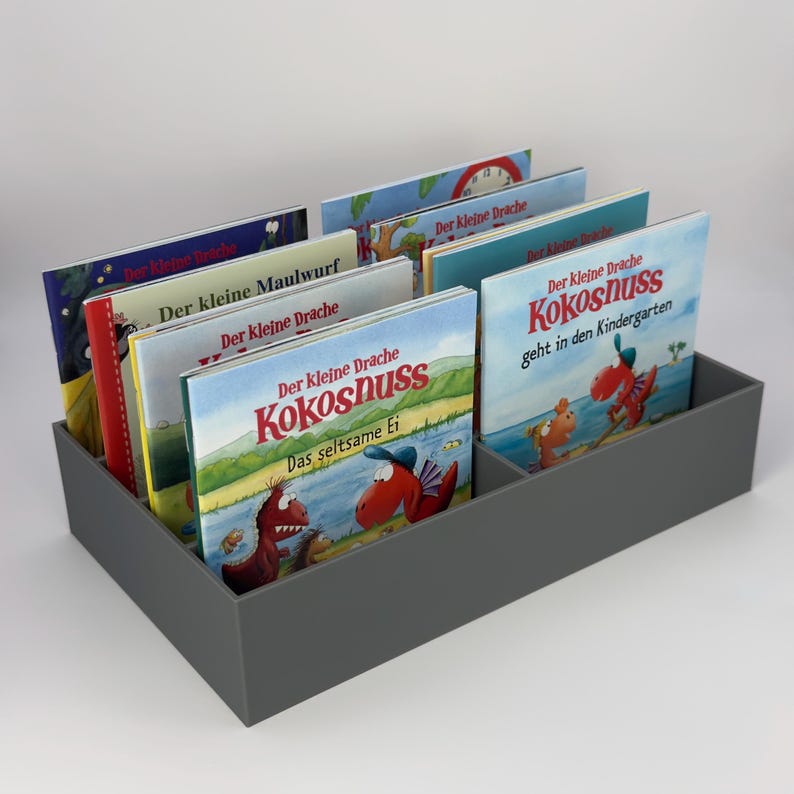 May include: A gray rectangular book organizer filled with children's books. The books have colorful covers with illustrations and German text, including "Der kleine Drache Kokosnuss" and "Der kleine Maulwurf". The organizer is divided into two sections.