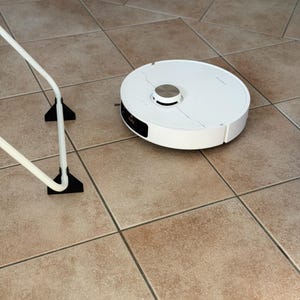 Könnte beinhalten: Ein weißer Roboterstaubsauger mit silbernem Akzent oben steht auf einem gefliesten Boden. Der Staubsauger hat ein kreisförmiges Design mit einem schwarzen Display. Ein weißes Metallobjekt mit schwarzen Stützen befindet sich im Vordergrund.