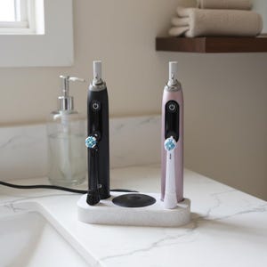 Könnte beinhalten: Zwei elektrische Zahnbürsten, eine schwarze und eine rosa, stehen in einer gesprenkelten weißen Ladestation. Die Zahnbürsten haben weiße Bürstenköpfe. Die Szene spielt in einem Badezimmer, im Hintergrund ist ein Seifenspender zu sehen.