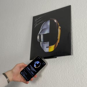 Könnte beinhalten: Schwarzes Vinyl-Schallplattencover mit geteiltem Helmdesign in Silber und Gold, mit dem Text "Random Access Memories". Ein Smartphone zeigt das Albumcover. Die Schallplatte ist an einer weißen Wand montiert.