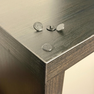 Könnte beinhalten: Nahaufnahme von vier kleinen, silberfarbenen Möbelprotektoren. Drei sind rund mit strukturierten Oberflächen, und einer hat einen Stiel. Sie sind auf der Ecke eines dunkelbraunen Holztisches platziert.