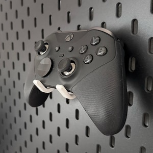 Könnte beinhalten: Ein schwarzer Xbox-Controller mit weißen Knöpfen und einer weißen Halterung. Der Controller ist an einer schwarzen Lochwand montiert.