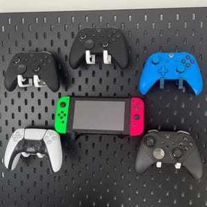Könnte beinhalten: Eine schwarze Lochwand mit weißen und dunkelgrauen Montagehaken, an denen vier Gamecontroller und eine Handheld-Spielkonsole befestigt sind. Die Controller umfassen zwei schwarze, ein blaues und ein weißes Modell, während die Konsole neon-grüne und pinke Seiten-Controller hat.