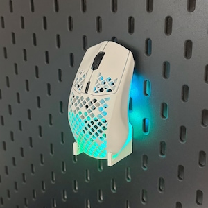 Könnte beinhalten: Eine weiße Gaming-Maus mit Wabenmuster und schwarzem Scrollrad ist auf einem weißen Ständer montiert. Die Maus hat blaue und grüne LED-Beleuchtung. Die Maus ist auf einer dunkelgrauen Lochwand montiert.