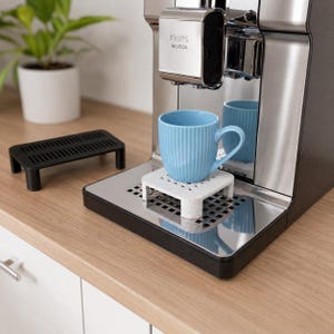 Könnte beinhalten: Ein hellblauer, gerippter Keramikbecher steht auf einem gesprenkelten, weißen, quadratischen Erhöhungselement mit vier kurzen Beinen auf der Abtropfschale einer Krups Intuition Kaffeemaschine aus Edelstahl. Ein schwarzer Kunststoffständer steht daneben.