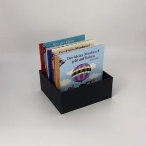 May include: A black rectangular box holding several children's books. The book covers feature colorful illustrations and German text. One book shows a hot air balloon. The books are stacked inside the box.