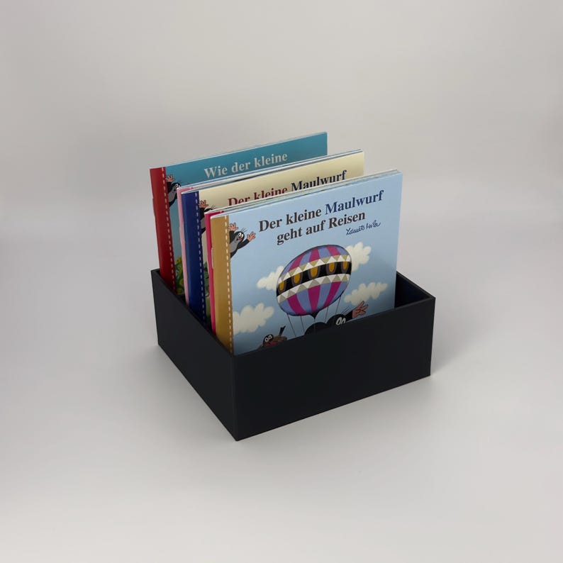 May include: A black rectangular box holding several children's books. The book covers feature colorful illustrations and German text. One book shows a hot air balloon. The books are stacked inside the box.