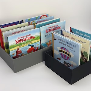 Könnte beinhalten: Zwei rechteckige graue und schwarze Buchorganisatoren, gefüllt mit Kinderbüchern. Die Bücher haben farbenfrohe Einbände mit deutschem Text und Illustrationen von Drachen, einem Maulwurf und einer Kokosnuss. Die Bücher sind aufrecht angeordnet.