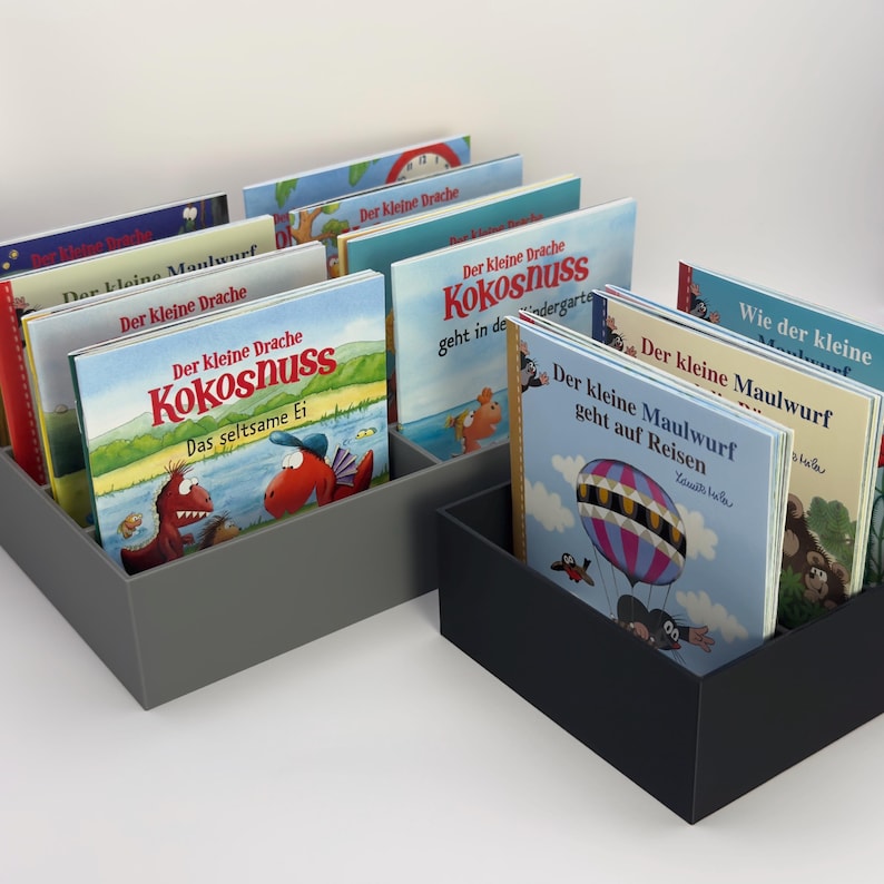 May include: Two rectangular gray and black book organizers filled with children's books. The books have colorful covers with German text and illustrations of dragons, a mole, and a coconut. The books are arranged upright.