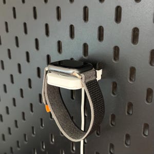 Könnte beinhalten: Eine silberne Smartwatch mit schwarzem Bildschirm und grau-schwarzem Stoffarmband ist auf einem weißen Ladeständer montiert. Die Uhr wird vor einem dunkelgrauen Lochwandhintergrund angezeigt.