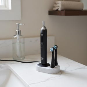 Könnte beinhalten: Eine schwarze elektrische Zahnbürste mit Ladestation und zwei Bürstenköpfen. Die Zahnbürste hat einen Ein-/Ausschalter und andere Bedienelemente. Die Bürstenköpfe haben blaue und weiße Borsten. Die Basis ist grau gesprenkelt.