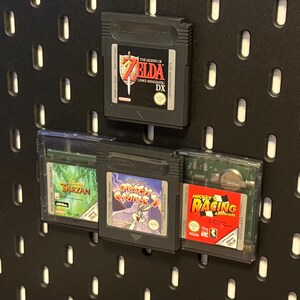 Könnte beinhalten: Drei Game Boy Color-Spielekartuschen, die auf einer schwarzen Lochwand ausgestellt sind. Die Kartuschen sind für die Spiele "The Legend of Zelda: Link's Awakening DX", "Disney's Tarzan", "Bugs Bunny: Crazy Castle 3" und "Mickey's Racing Adventure".