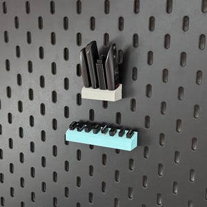 Könnte beinhalten: Ein weißer Wandorganizer mit vier schwarzen elektronischen Geräten und ein hellblauer Wandorganizer mit acht kleinen schwarzen USB-Empfängern, beide an einer dunkelgrauen Lochplatte mit vertikalen ovalen Löchern befestigt.