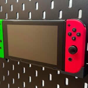 Könnte beinhalten: Eine tragbare Videospielkonsole mit schwarzem Bildschirm, einem neongrünen linken Controller und einem neonroten rechten Controller. Die Konsole ist horizontal in einer schwarzen Kunststoffhalterung an einer schwarzen Lochwand befestigt.