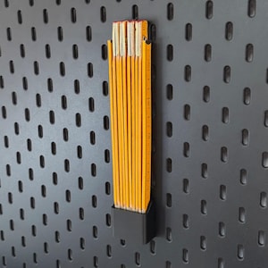 Könnte beinhalten: Ein gelb-oranges Zollstock, gehalten in einem schwarzen Halter, vor einer dunkelgrauen Lochwand. Das Maßband ist mit Zentimetern markiert. Ein Werkzeug für die Zimmerei.