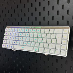 Könnte beinhalten: Weiße Gaming-Tastatur mit mehrfarbig beleuchteten Tasten. Die Tastatur hat ein kompaktes Design mit dem Text "REPUBLIC OF GAMERS" oben. Die Tasten sind in einem Standard-QWERTZ-Layout angeordnet.