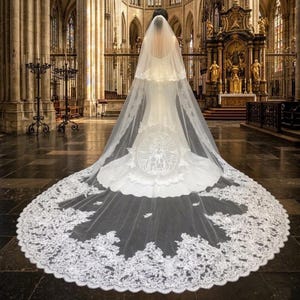 Op de afbeelding: Een vloerlange witte trouwjurk met een lange, doorschijnende kathedraalsluier met ingewikkelde kanten randen. De achterkant van de jurk toont een cirkelvormig geborduurd ontwerp van een religieuze figuur, waarbij de sluier over de vloer uitwaaiert.
