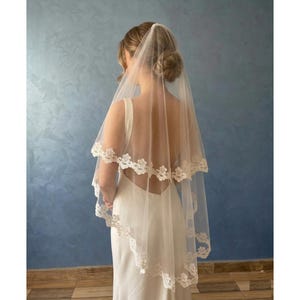 Pode incluir: Um véu de noiva marfim de duas camadas com delicada renda floral. O véu é transparente, permitindo que o vestido seja visível. A noiva está usando um vestido branco com as costas abertas e o cabelo está preso em um coque.